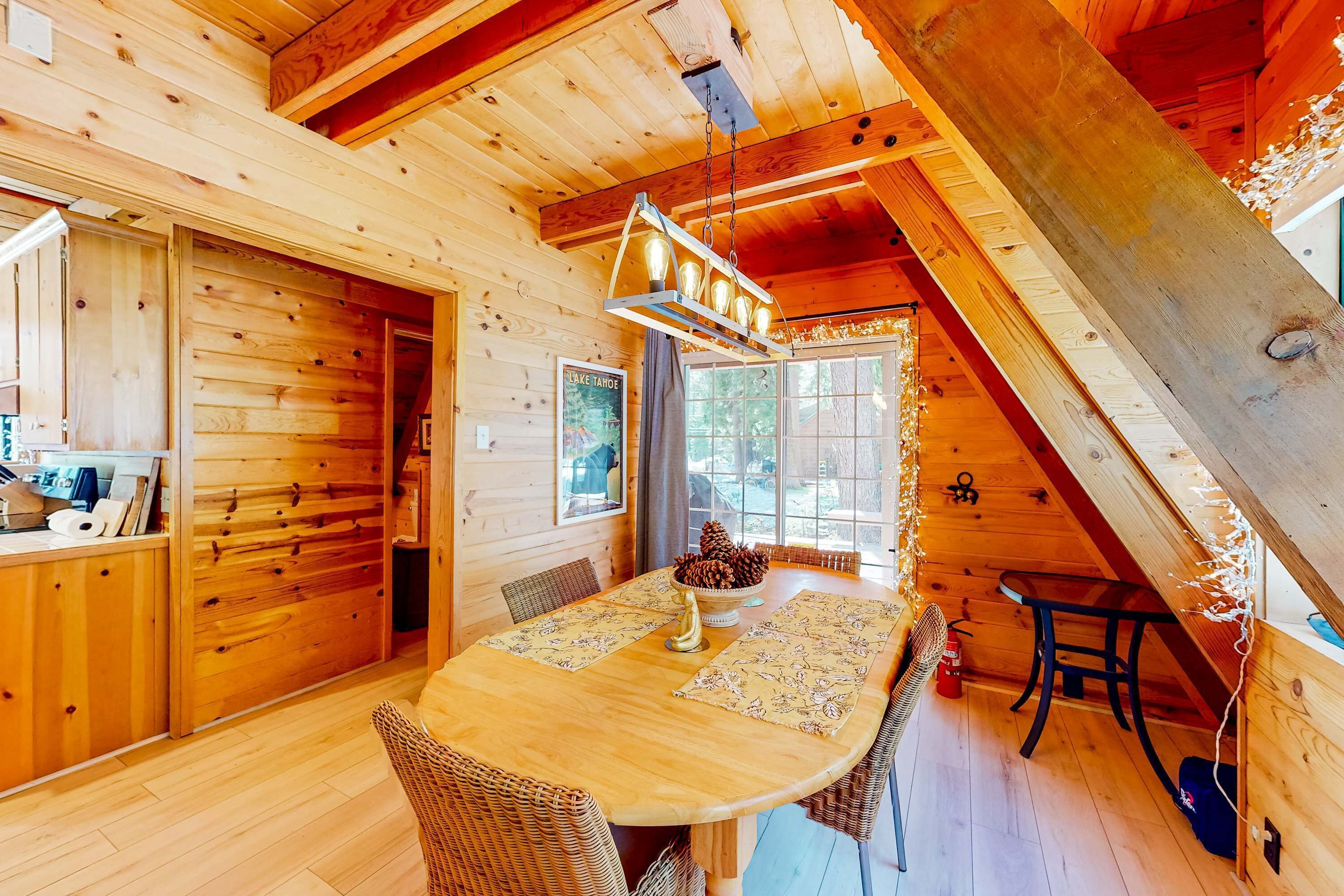 Interior de una cabaña de madera con una mesa de comedor, sillas, una lámpara colgante y una ventana con vistas.