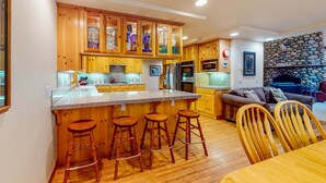 A kitchen with wooden cabinets, a central island with stools, a stone fireplace, and a dining area with wooden chairs. - Mountain Home with Access to Shared Pool, Hot Tub, Sauna, & Tennis (Truckee)