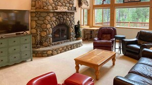 A living room with a stone fireplace, leather sofas, a television, and a view of a lake. - Mountain Home with Access to Shared Pool, Hot Tub, Sauna, & Tennis (Truckee)