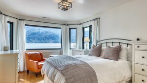 A bedroom with a large bed, a desk, and a chair. There are multiple windows with curtains, and a view of a forested area outside. - Dog-friendly lakefront home with fast WiFi, private dock, & 180-degree views (Truckee)