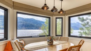 A dining area with a round table, wicker chairs, and large windows offering a view of a lake and mountains. - Dog-friendly lakefront home with fast WiFi, private dock, & 180-degree views (Truckee)
