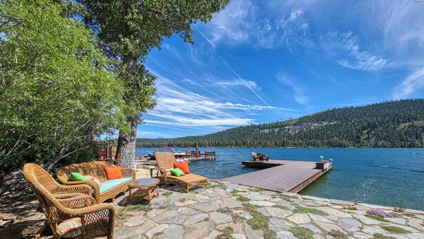 A lakeside patio with wicker furniture, a dock extending into the water, and a forested hill in the background. - Dog-friendly lakefront home with fast WiFi, private dock, & 180-degree views (Truckee)