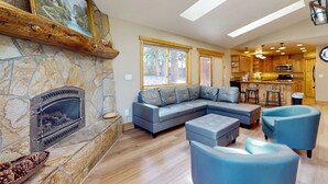 A living room with a stone fireplace, a gray sectional sofa, and a kitchen in the background. - Grand lodge in the woods near golf, hiking & skiing with multiple fireplaces (South Lake Tahoe)