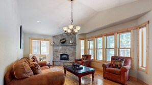 A spacious living room with a stone fireplace, wooden flooring, and large windows. - Grand lodge in the woods near golf, hiking & skiing with multiple fireplaces (South Lake Tahoe)