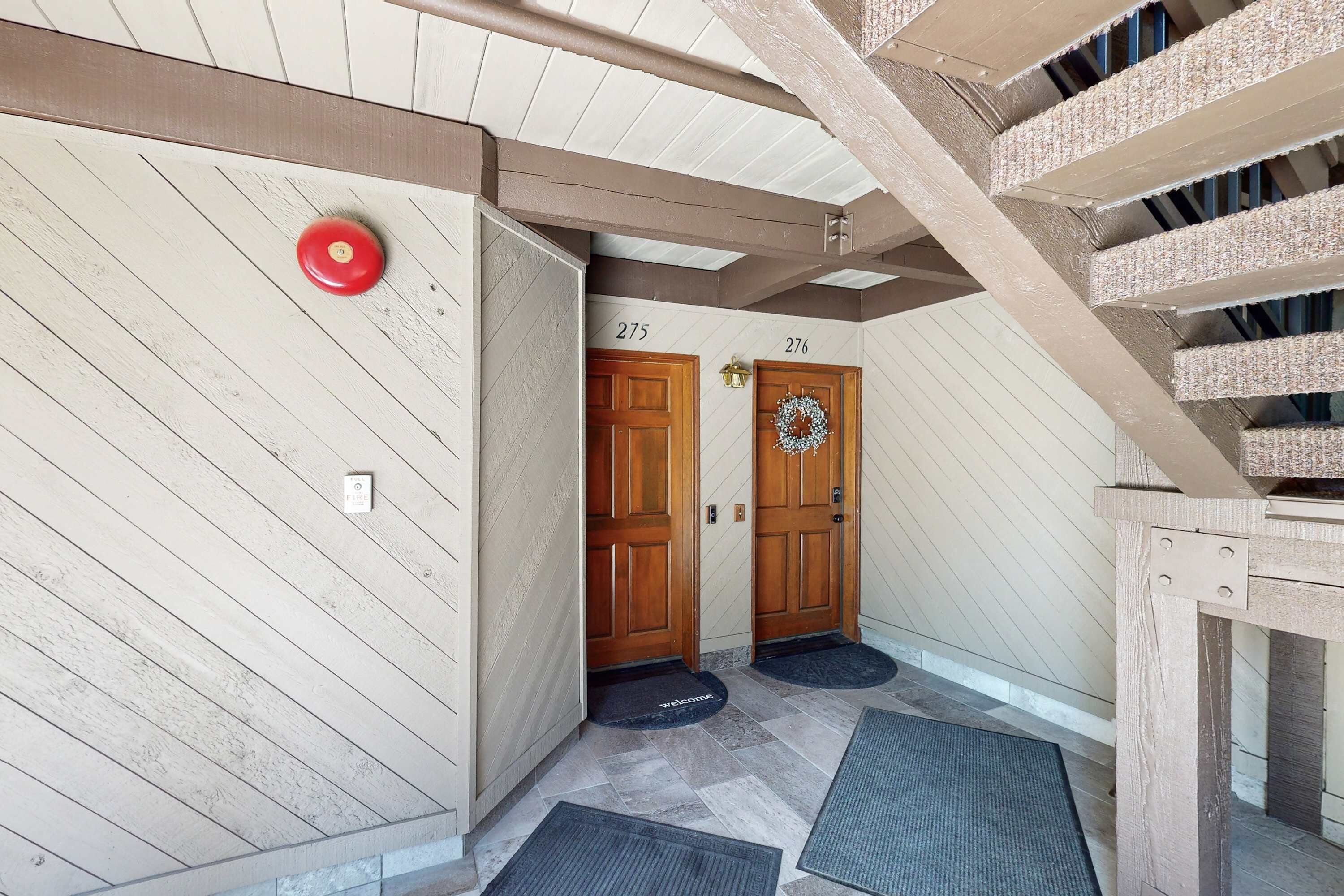 A hallway with wooden doors labeled 275 and 276, a red emergency button, and a welcome mat.
