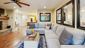 Ein Wohnzimmer mit einem Ecksofa, einem Couchtisch mit Blumen, einem Kamin und einem Deckenventilator.