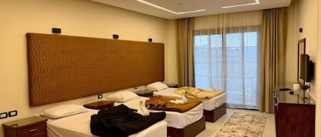 Habitación de hotel con una cama grande, dos camas más pequeñas, un cabecero de madera, una mesita de noche, un televisor y vistas a la ciudad desde la ventana.
