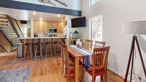 A modern kitchen with wooden cabinets, a central island, and a dining area with wooden chairs and stools.