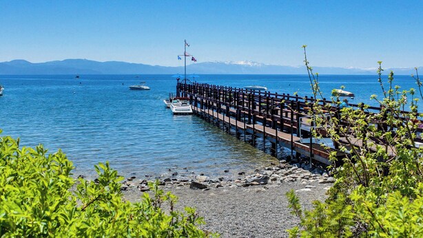 Un muelle se adentra en un lago tranquilo, con barcos amarrados, rodeado de montañas y cielos despejados.
