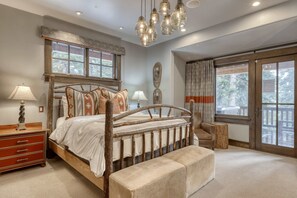 A bedroom with a wooden bed, bedside lamps, a bench, and a view of the outdoors through large windows.