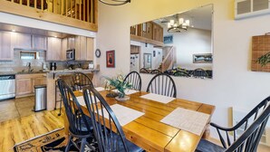 Uma ampla área de jantar com mesa e cadeiras de madeira, uma cozinha com armários de madeira, e um mezanino com área de dormir e guarda-corpo de madeira.