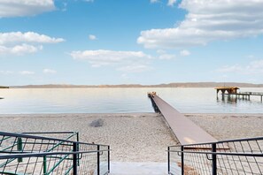 Ein Strand mit einer hölzernen Pier, die sich über ruhiges Wasser erstreckt, einem sandigen Ufer und einer entfernten Bergkette.