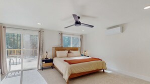 Una camera da letto con un letto, una ventola a soffitto, un comodino, una lampada e una vista su un balcone esterno attraverso una porta scorrevole.