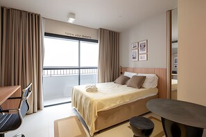 Quarto de hotel com cama, escrivaninha, cadeira, mesa pequena e varanda com cortinas.