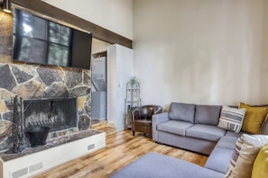 A living room with a stone fireplace, a grey sofa, and a flat-screen TV mounted above the fireplace. - Cozy and Convenient Condo with Fireplace, Deck, W&D, Close to Town, Lake, Trails (Tahoe City)