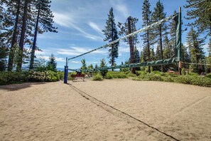 Une plage de sable avec un filet de volley-ball, entourée de grands arbres et sous un ciel dégagé.