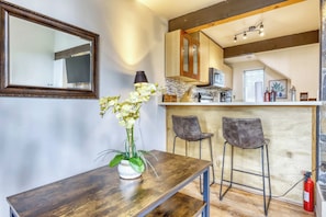 A dining area with a wooden table, two bar stools, a vase with flowers, a mirror, and a kitchen in the background. - Cozy and Convenient Condo with Fireplace, Deck, W&D, Close to Town, Lake, Trails (Tahoe City)