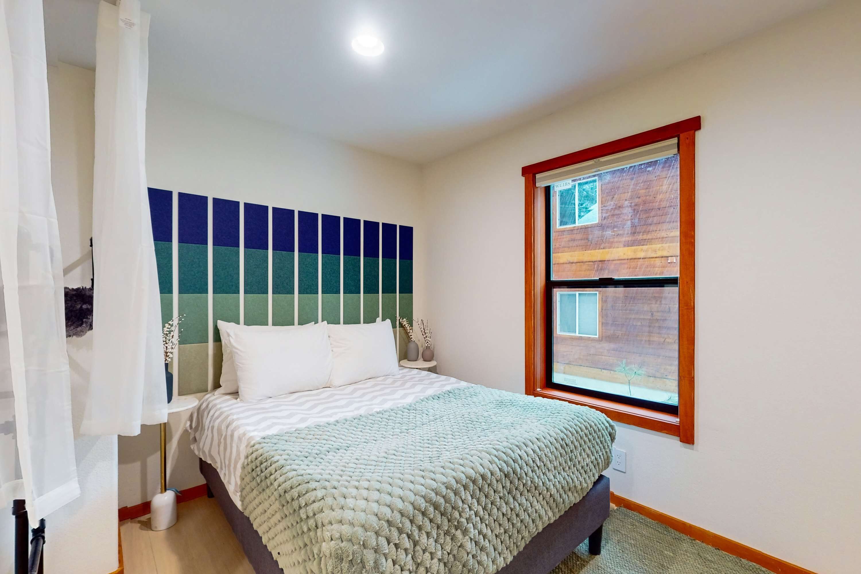 Un dormitorio con cama, ventana, pared con estampado azul y verde, y mesita de noche con un jarrón.