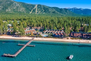 Een luchtbeeld van een kustresort met een pier, strandwoningen en een bergachtige achtergrond.