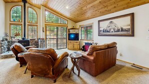 A spacious living room with leather sofas, a fireplace, a television, and large windows. - Charming mountain home with game room, decks, and stunning views (Truckee)