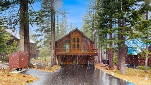 A wooden cabin with a covered porch, surrounded by tall trees and a clear sky. - Charming mountain home with game room, decks, and stunning views (Truckee)