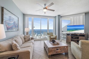 A spacious living room with a large flat-screen TV, a ceiling fan, a sofa, a coffee table, and a balcony with a view of the ocean. - Lighthouse 1615 by Liquid Life (Gulf Shores)
