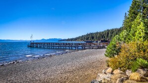 A rocky shoreline with a wooden pier extending into calm waters, surrounded by dense forest and distant mountains. - Homey Family Condo with Shared Pool, Hot Tub, Tennis & Lovely Lake Views (Tahoe City)