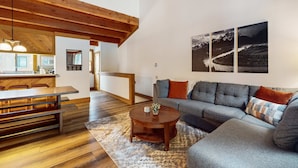 A modern living room with a wooden ceiling, a grey sofa, a round coffee table, and a dining area with a wooden table and benches. - Cozy townhouse with pool access, board games, and fireplace - near skiing (Northstar - Truckee)