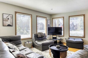 A living room with a grey sofa, a black leather chair, a TV on a stand, and large windows with blinds.