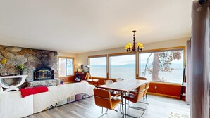 Uma sala de estar ampla com lareira de pedra, mesa de jantar com cadeiras e grandes janelas com vista para o mar.