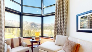 Ein Zimmer mit großem Fenster und Blick auf die Berge, eine Couch, ein Sessel und ein kleiner Tisch mit Lampe.