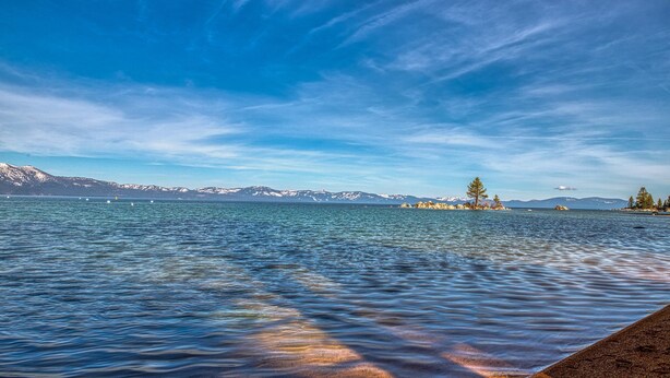 Ein ruhiger See mit klarem Wasser, fernen Bergen und einem wolkenlosen Himmel.