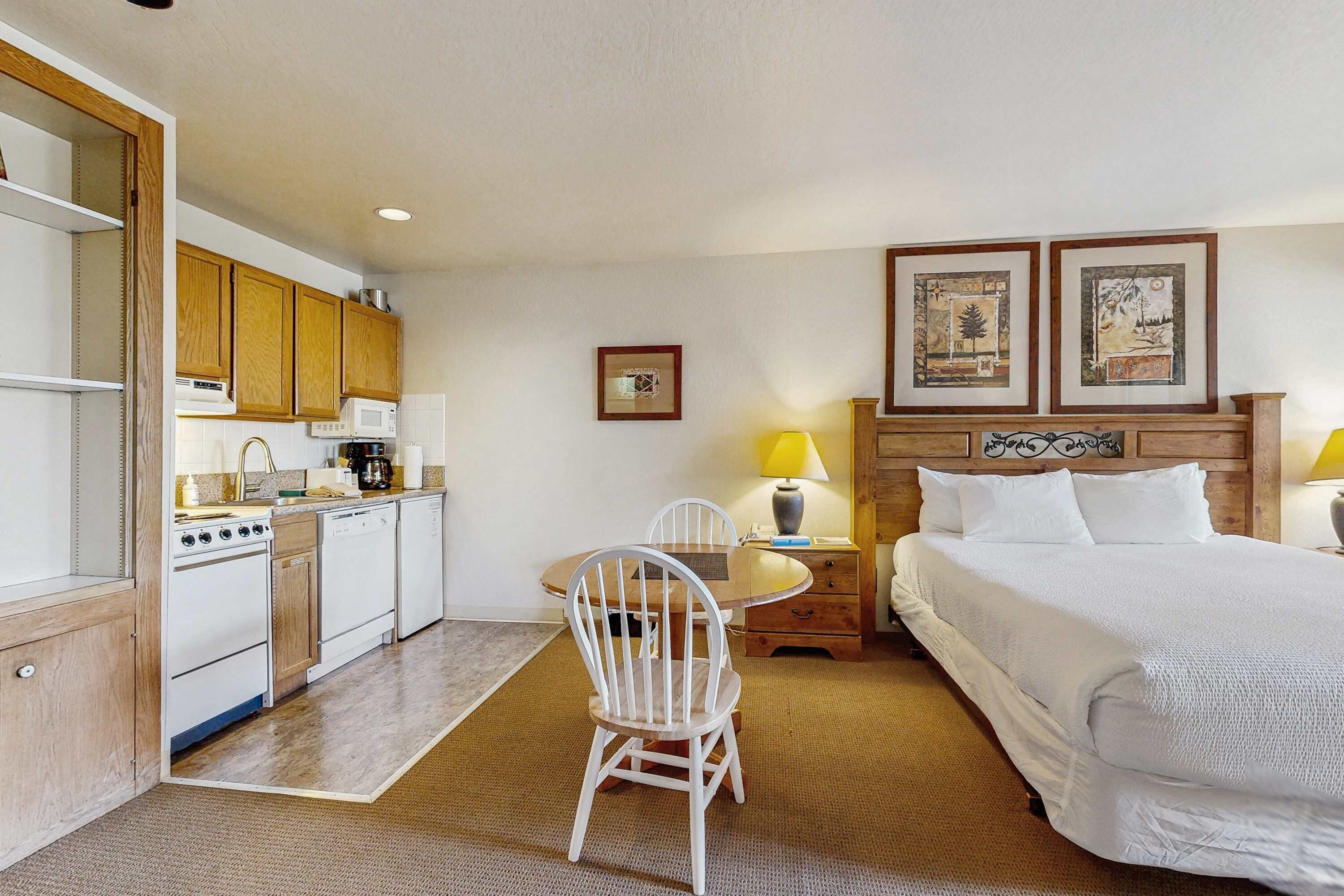 Habitación de hotel con cama, mesa de comedor con sillas, zona de cocina y obras de arte en las paredes.