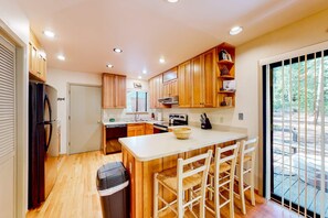 Une cuisine avec des meubles en bois, un îlot central doté de chaises, une poubelle et une vue sur l’extérieur grâce à une porte vitrée.