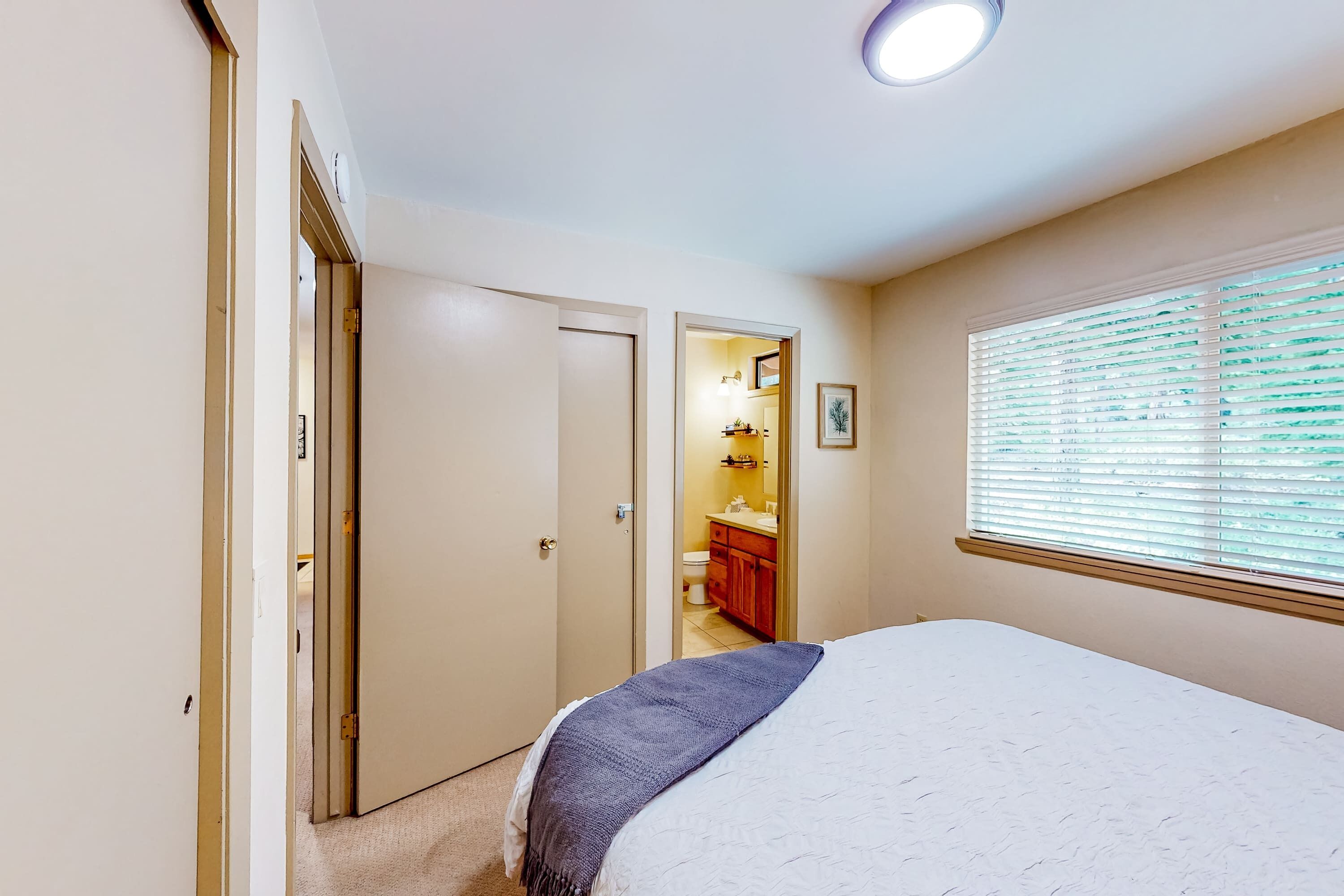 Quarto com cama, janela com persianas, porta para um banheiro e luminária no teto.