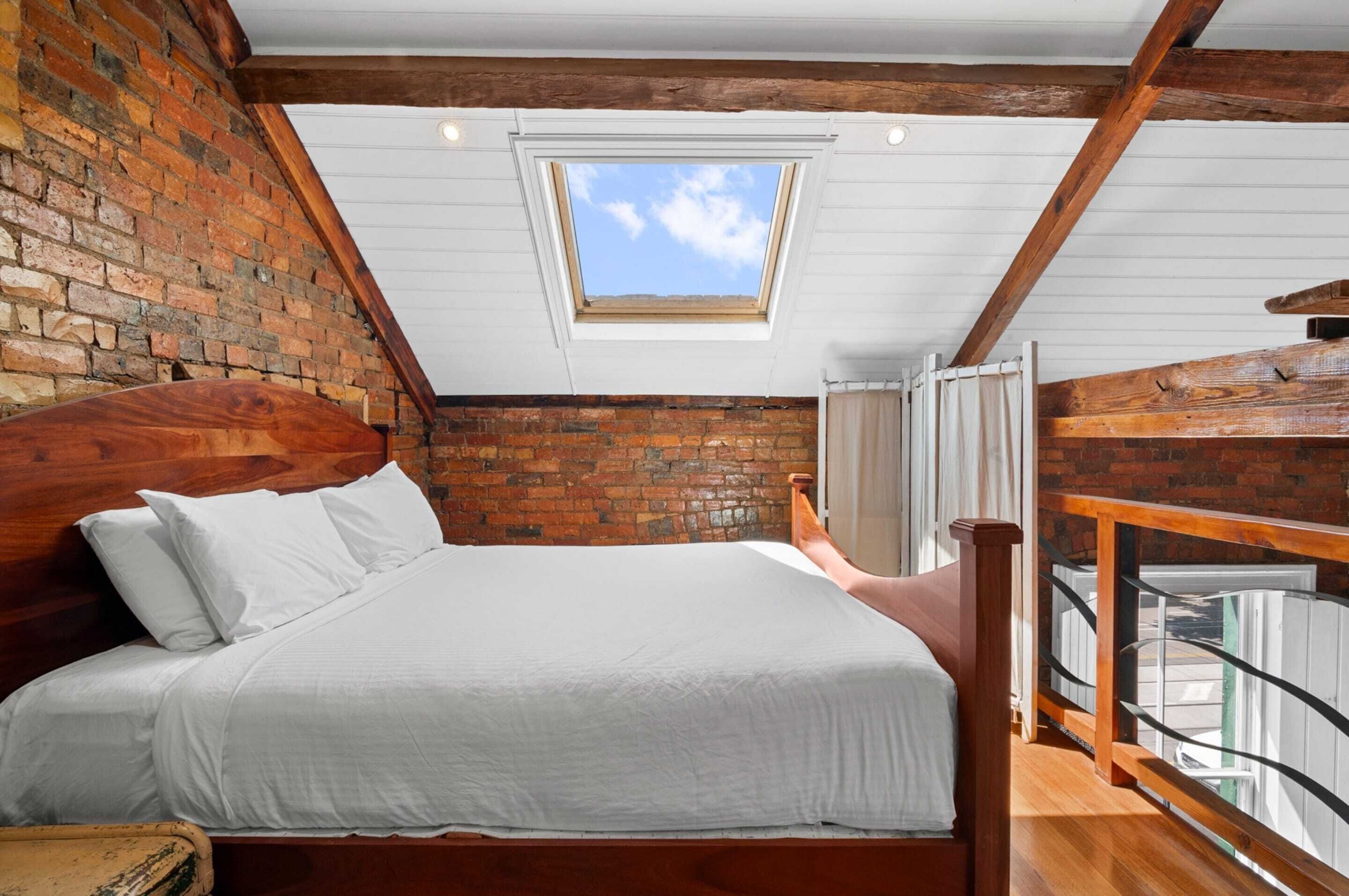 Een slaapkamer met schuine balken, een dakraam, een houten bed, een bakstenen muur en een balkon met een hekwerk.