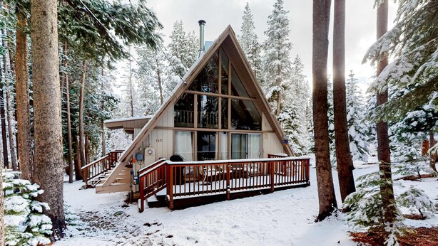 Een chalet met een houten terras, een trap en een groot raam, omringd door met sneeuw bedekte bomen.