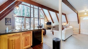 Een ruime loft met keuken, eethoek en zithoek, met houten balken, grote ramen en uitzicht op bomen.
