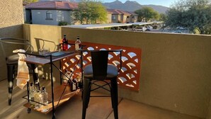 En balkon med et bord, stole og flasker, med udsigt over et boligområde med huse og bjerge.