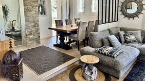 A living room with a stone accent wall, a dining area with a wooden table and chairs, a grey sofa with patterned cushions, and a decorative mirror with a sunburst frame. - Luxury 5 BR Villa with Pool in St Paul-de-Vence (Saint-Paul-de-Vence)