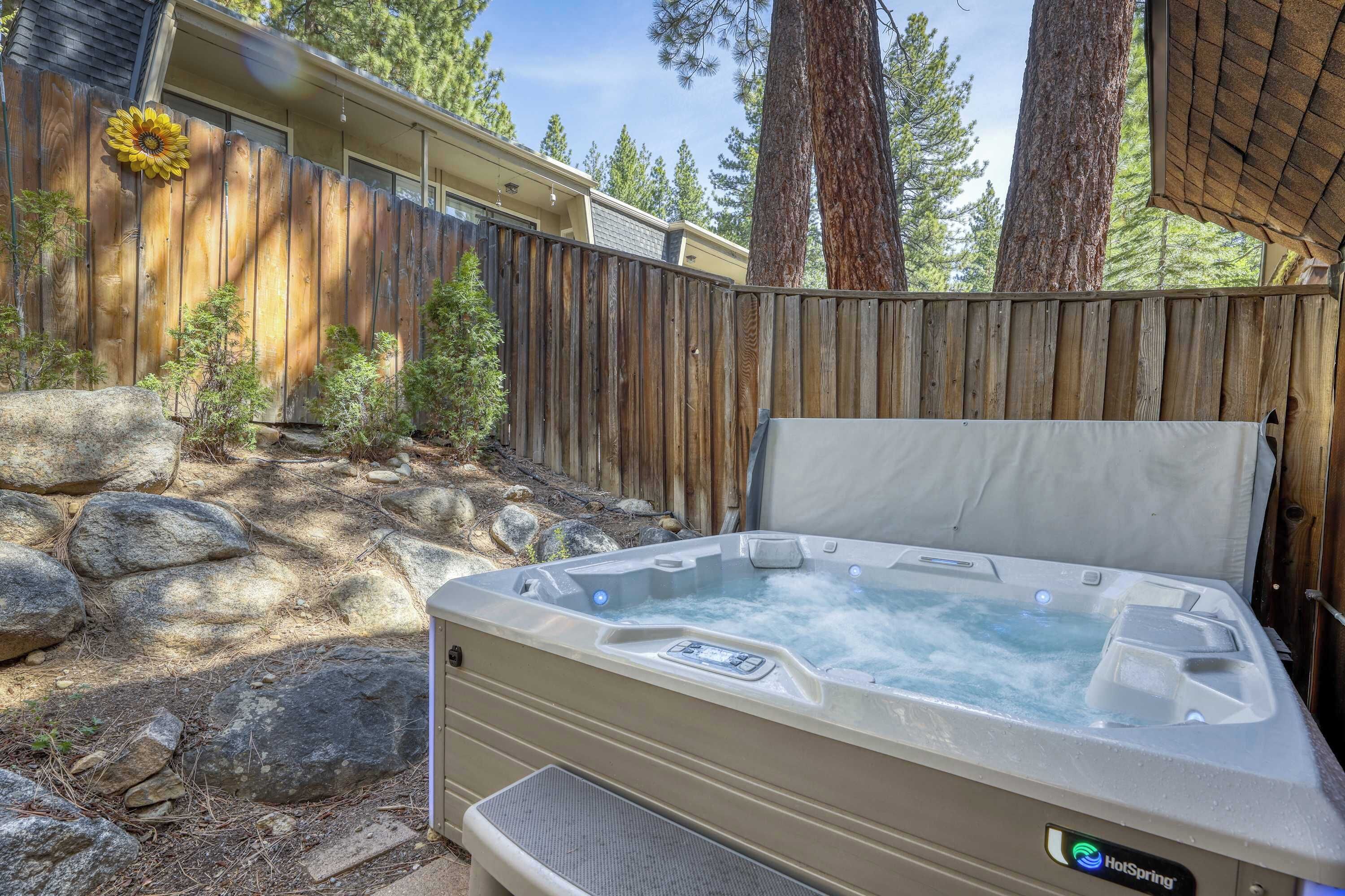 A hot tub in an outdoor setting with a wooden fence, rocks, and trees in the background.