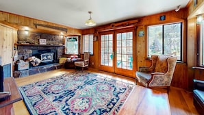 A log cabin interior with a stone fireplace, wooden walls, a large rug, and a view through the glass doors. - Riverfront dog-friendly retreat with a rustic flair & wood stove, near skiing (Soda Springs)