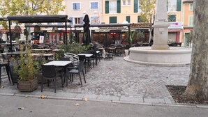Un área de café al aire libre con mesas y sillas, una fuente y edificios al fondo.