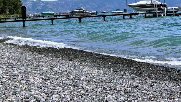 Una playa pedregosa con barcos amarrados en un muelle, montañas al fondo y un cielo despejado.