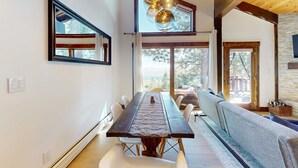 Un espace repas avec une longue table, des chaises blanches et un chemin de table à motifs. On y trouve des luminaires sphériques suspendus et, au fond, un mur en pierre.