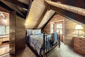 Een houten slaapkamer met schuine wanden, een bed met beddengoed met een patroon, een dressoir en een lamp.