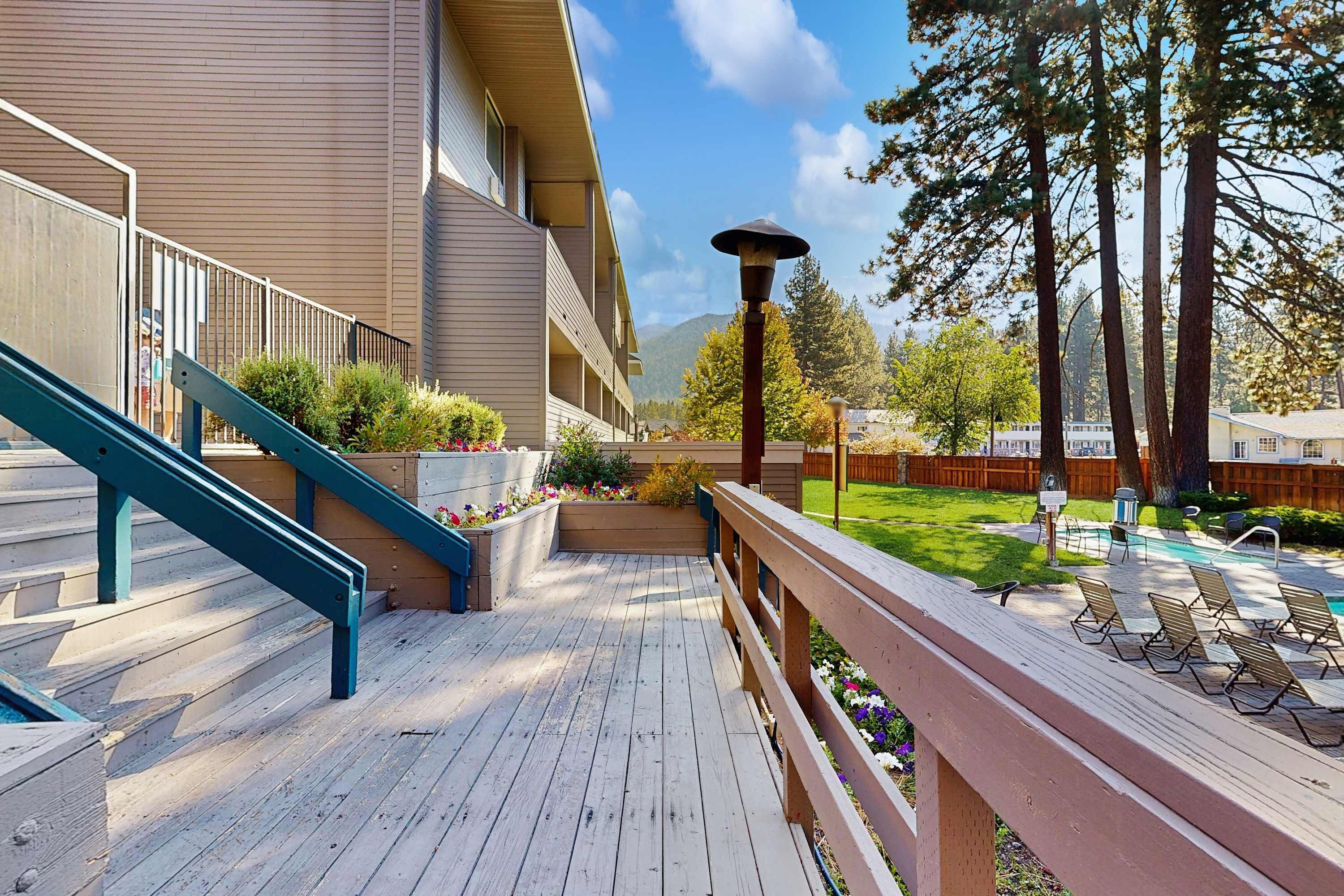 Una terraza de madera con una escalera azul, un área de piscina con sillones de descanso y un edificio con balcón.