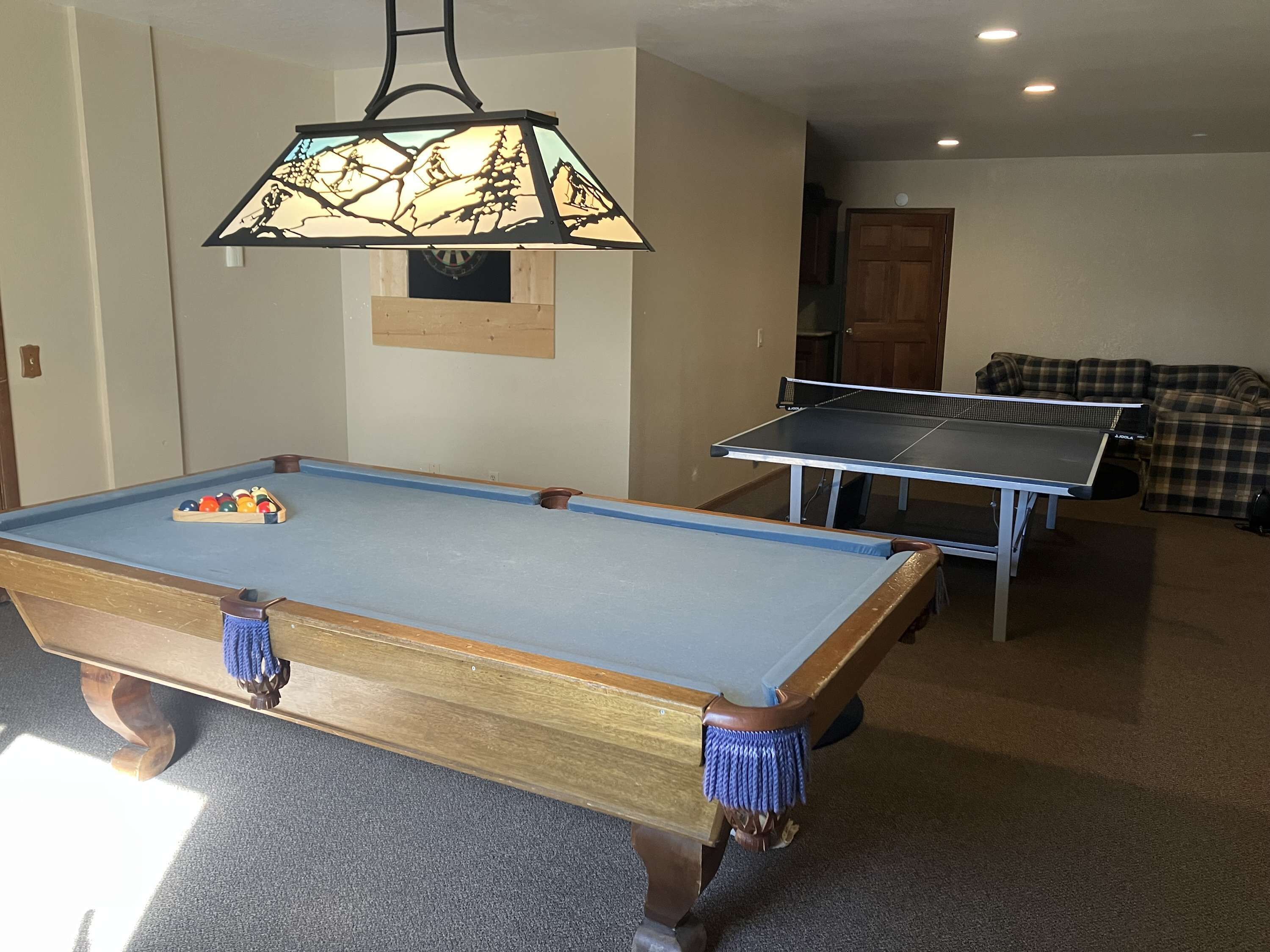 Une table de billard recouverte d’un drap bleu, un porte-queues et une lampe suspendue représentant un paysage de montagne.