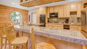 Una cocina con armarios de madera, encimeras de granito, una isla central con asientos y un ventanal con vista a árboles.