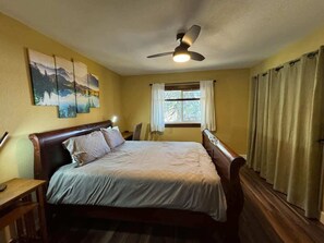 Un dormitorio con cama, ventilador de techo, ventana con cortinas y un cuadro en la pared.